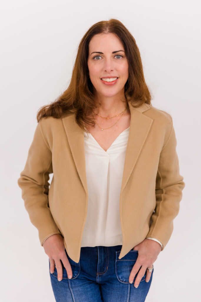 woman standing in front of white background arms crossed wearing a white top, beige jacket and jeans during headshot photoshoot with Laure Photography