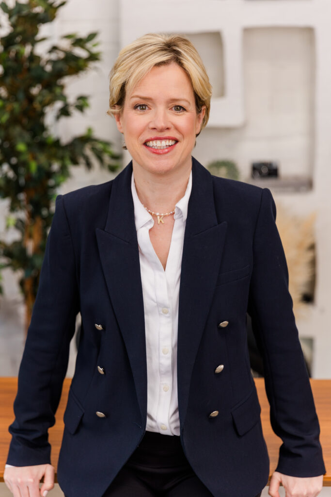 woman leaning against a desk one hand on each side with a lifestyle office background wearing corporate outfit white shirt and dark blue jacket during headshot photoshoot with Laure Photography