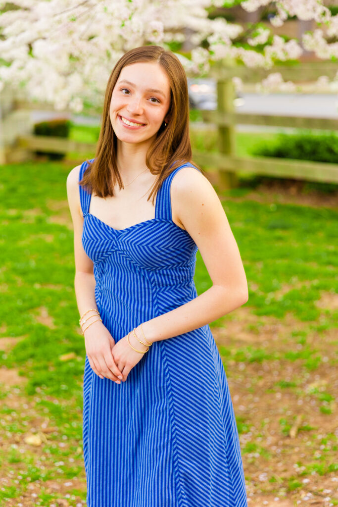 High school girl wearing a long sleeveless blue dress with stripes holding hands together in front of a blooming tree in a park and smiling at the camera during senior portraits session in Atlanta GA
