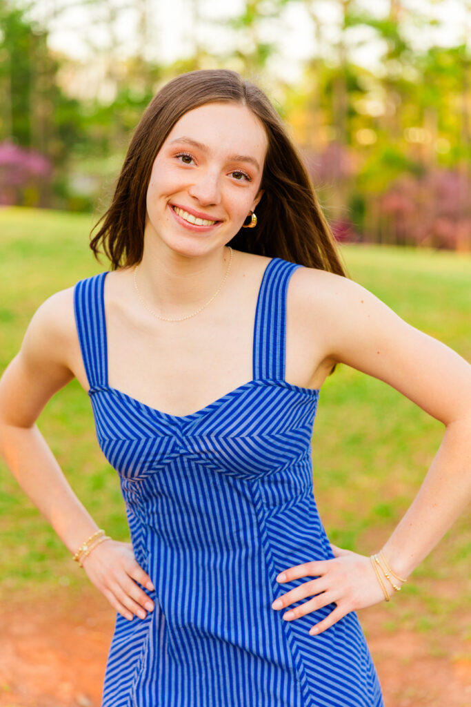 senior girl wearing a long sleeveless blue dress with stripes standing in a park both hands on her hips during portraits session in Atlanta GA