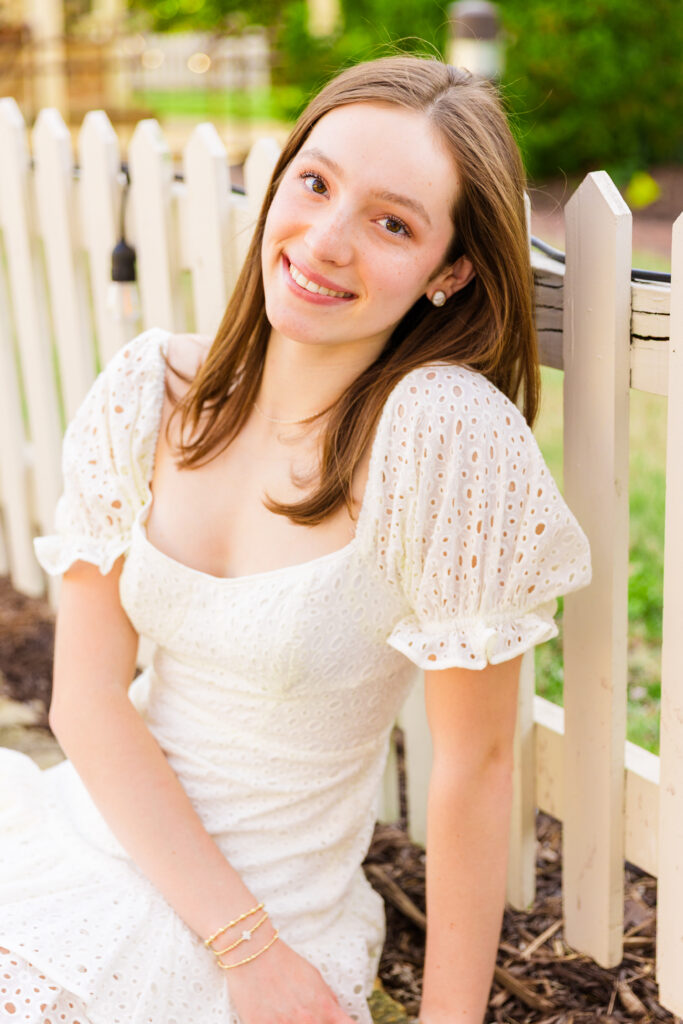 senior girl wearing a short textured white dress sitting on front of a white fence during portraits session in Atlanta GA