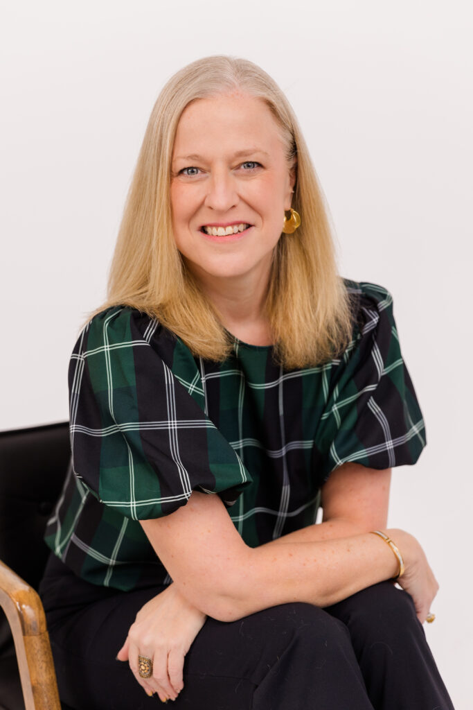 woman with blond hair sitting on a chair in front of white background wearing corporate outfit tartan fabric top and black pant during headshot photoshoot with Laure Photography