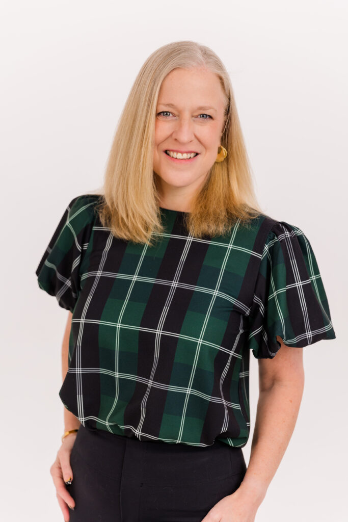 woman with blond hair standing in front of white background wearing corporate outfit tartan fabric top and black pant during headshot photoshoot with Laure Photography