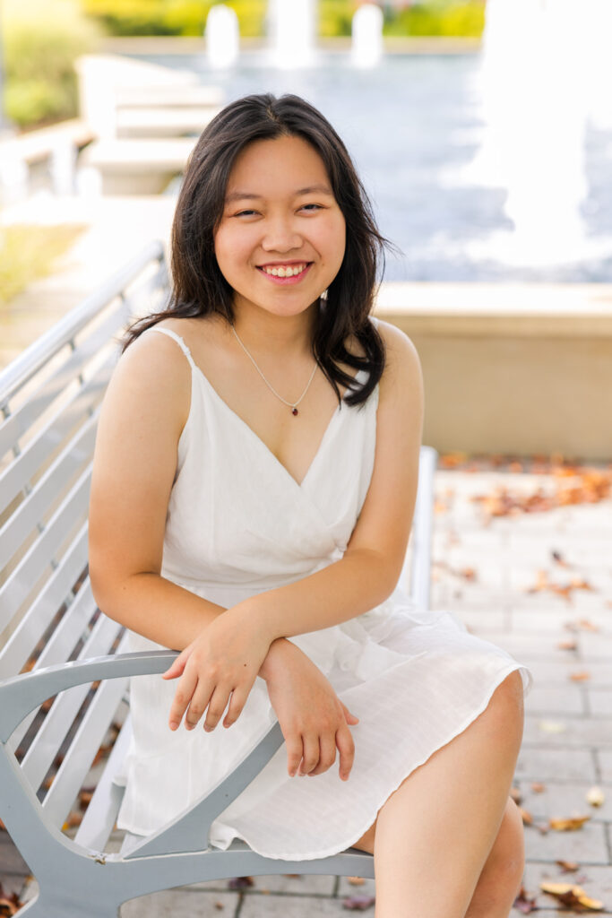 High school senior girl in white dress sitting on a bench in front of a fountain during her joyful senior portraits with Laure Photography.
