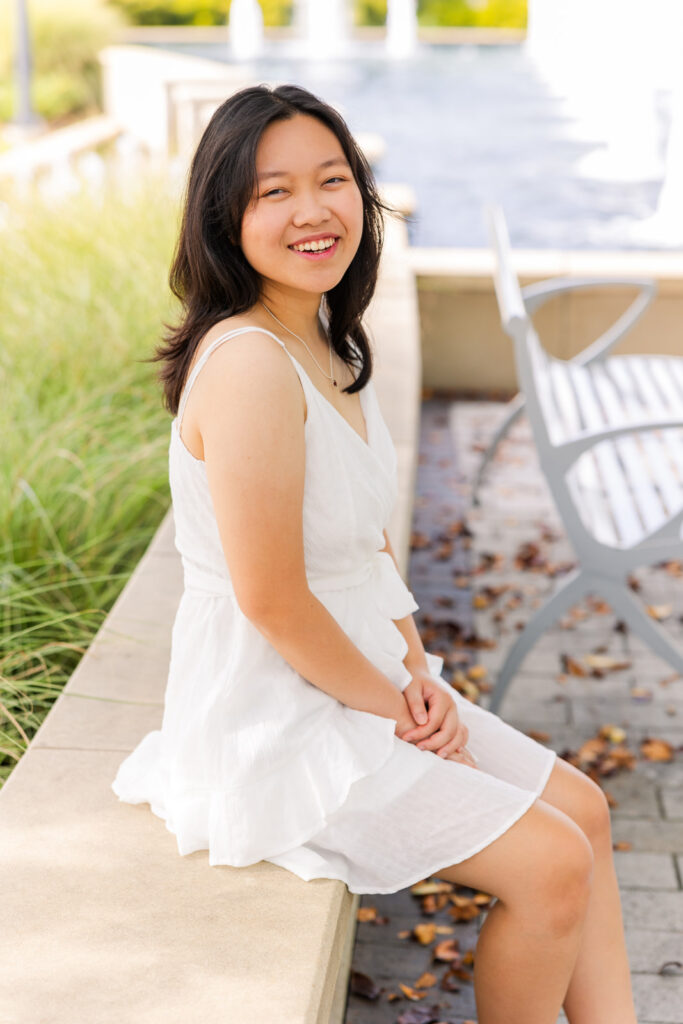 Senior girl in white summer dress sitting on the wall by the fountain during her joyful Sandy Springs senior photoshoot with Laure Photography.