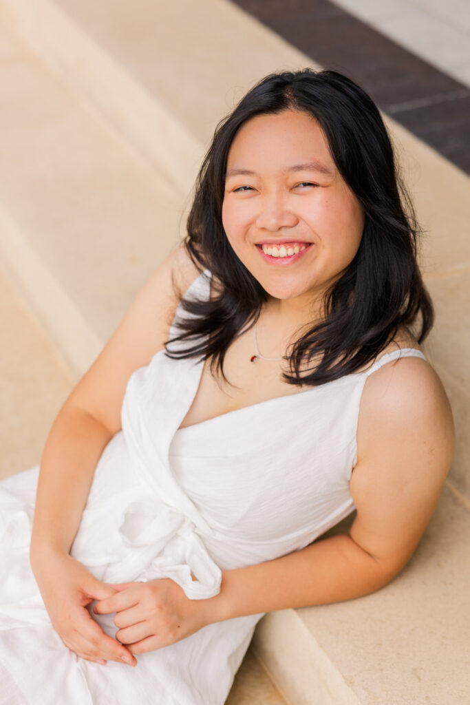 Senior girl in white sleeveless dress sitting on outdoor stairs, leaning on her elbow during portraits with Laure Photography in Sandy Springs.