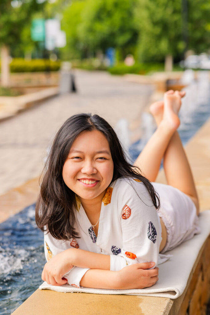 Senior girl wearing patterned shirt and shorts laying on her elbows by a fountain during her Sandy Springs senior session with Laure Photography.