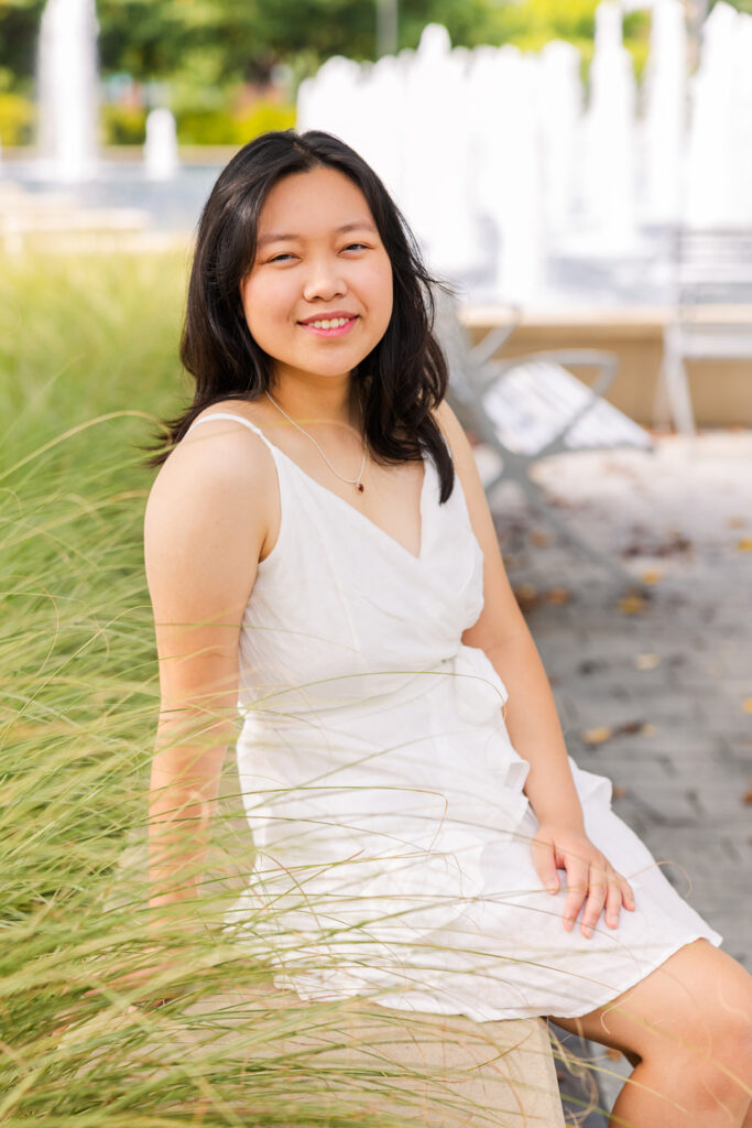 High school senior girl in white summer dress sitting on a wall in front of a fountain during her Atlanta senior photoshoot with Laure Photography.