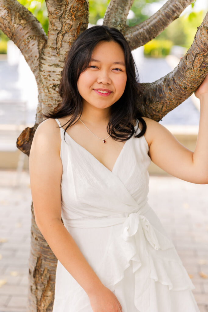 Senior girl leaning against a tree, holding a branch, wearing a white sleeveless dress during her Sandy Springs portraits with Laure Photography.