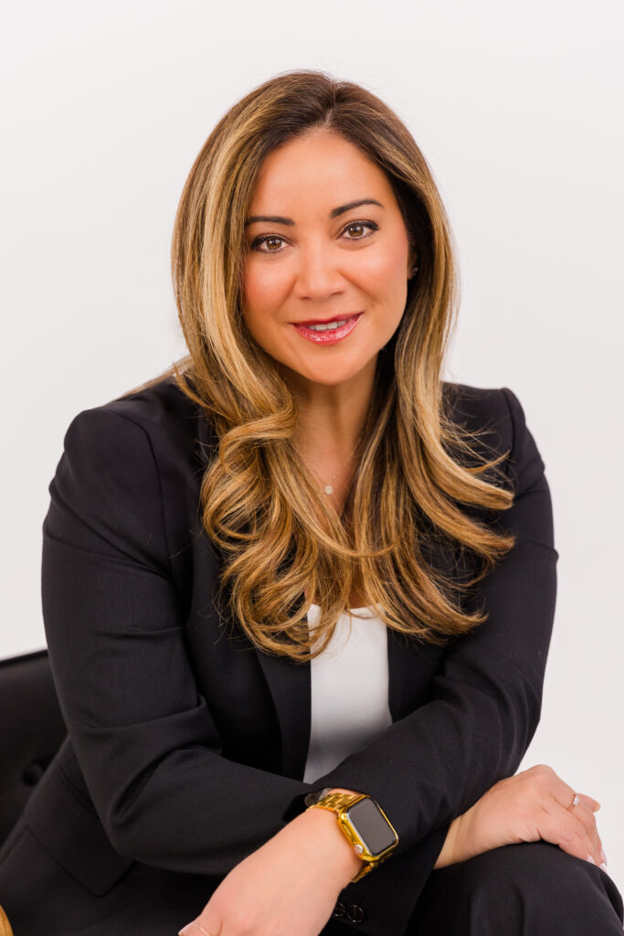 woman with long hair sitting on a chair in front of white background wearing corporate outfit white top and dark jacket during headshot photoshoot with Laure Photography
