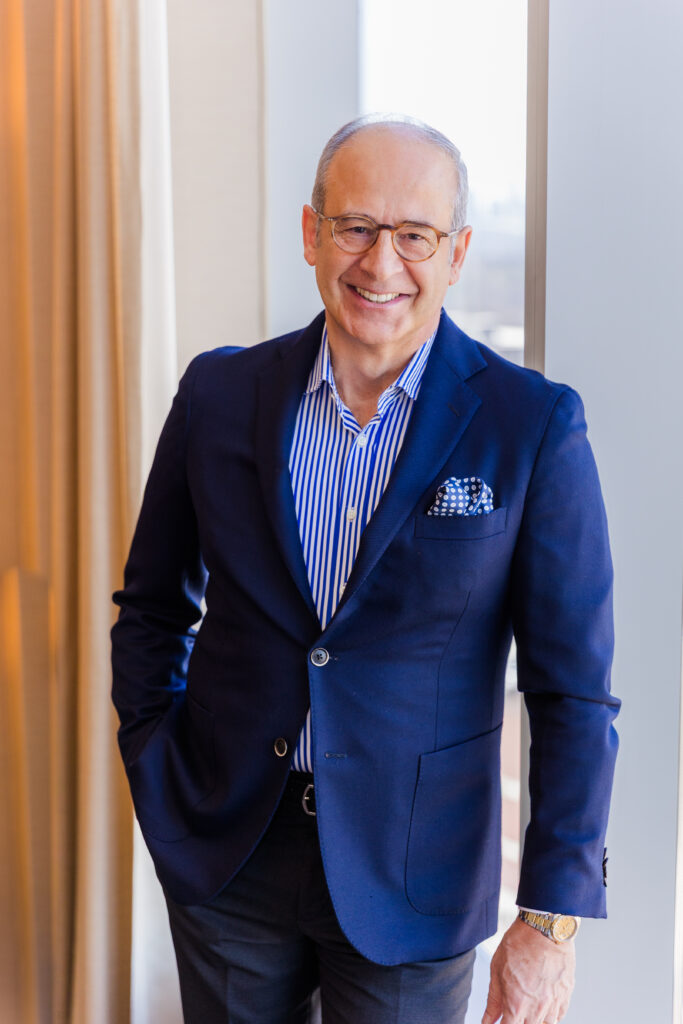 Ahmet Bozer wearing a blue and white striped shirt, grey pants, and navy jacket standing against a window, hand in pocket, looking at the camera