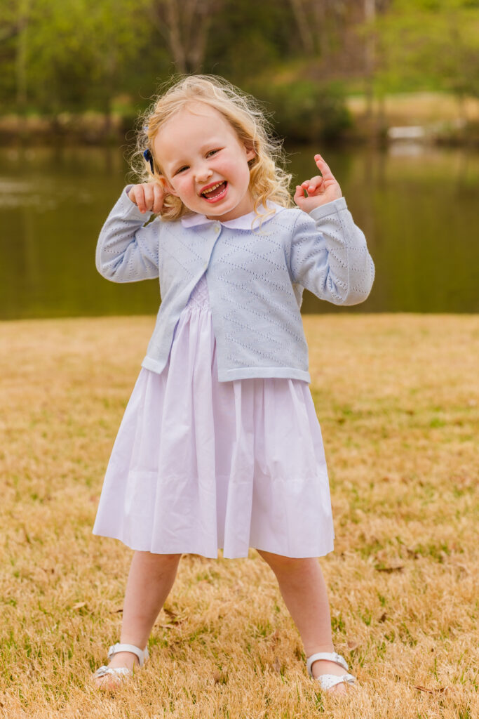 Toddler girl in a white dress and blue jacket smiling brightly during a family photo session in an Atlanta park by Laure Photography