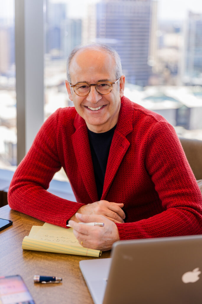 Ahmet Bozer wearing a red casual jacket and black t-shirt, taking notes at his desk in front of large windows with an Atlanta city skyline view