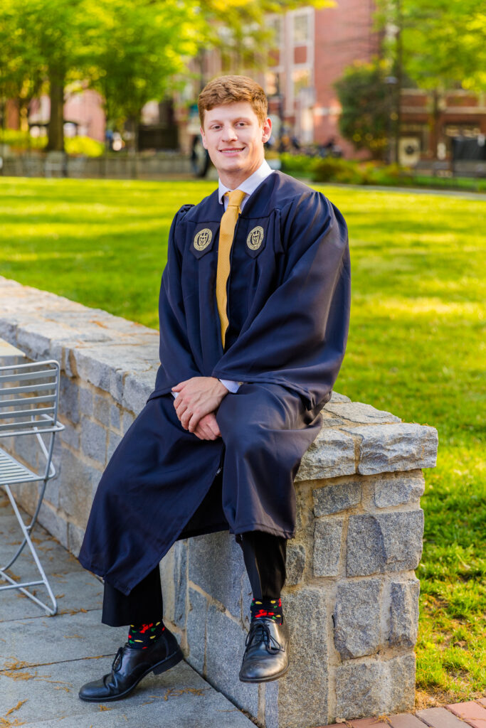 Jason sitting on a low brick wall outdoors in his graduation outfit.