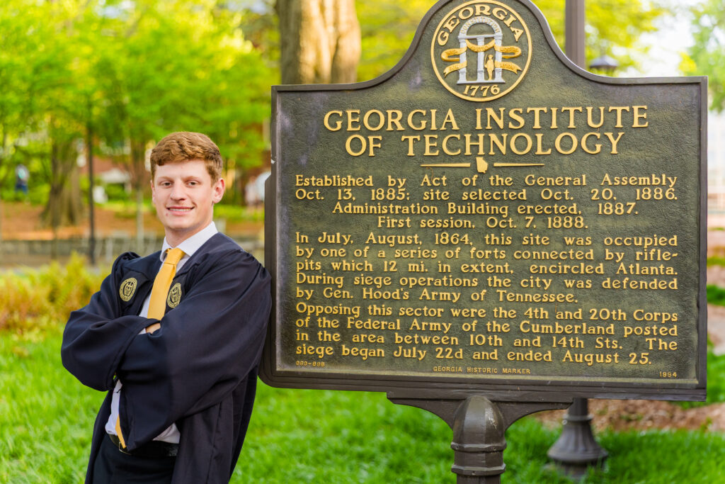 closeup Jason standing by the Georgia Tech campus history sign in graduation gown leaning on the sign arms crossed