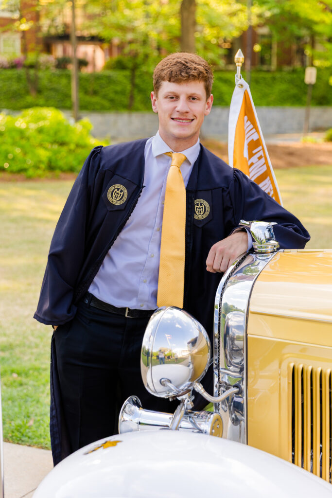 Jason leaning on the front of the Ramblin' Wreck wearing his cap and gown.