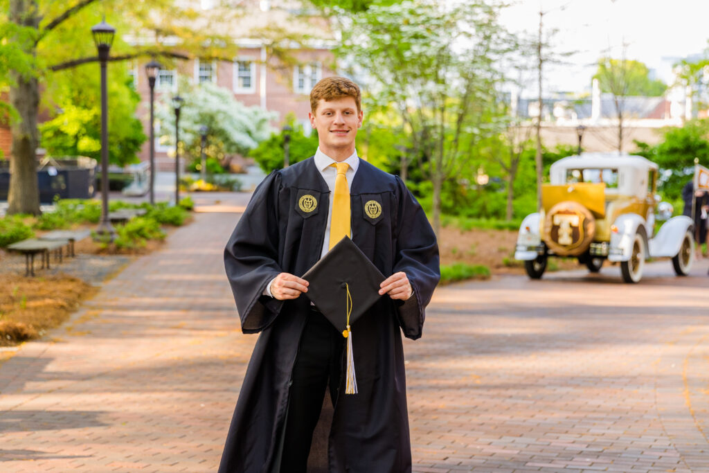 Jason standing with his graduation hat in hand, Ramblin' Wreck and campus behind him.
