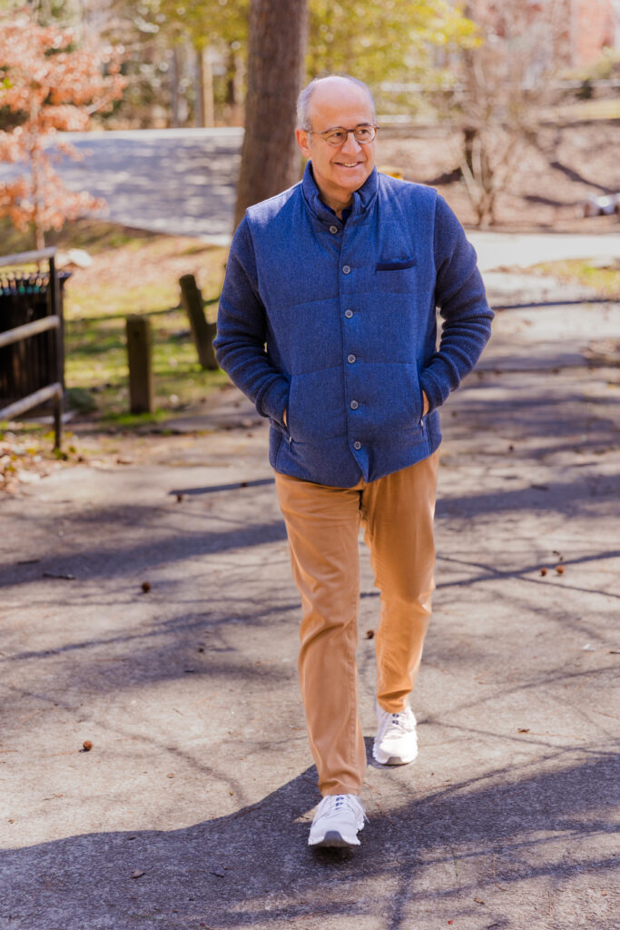 Ahmet Bozer walking hands in pockets through an Atlanta Buckhead park wearing a sleeveless jacket, beige pants, and walking shoes