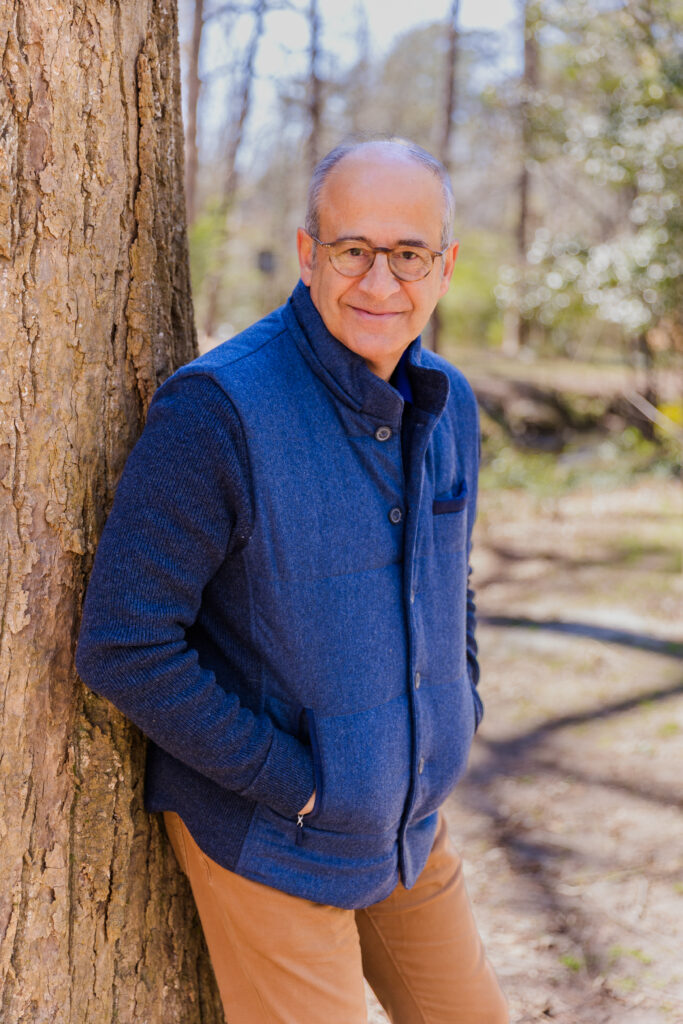 Ahmet Bozer leaning against a tree, hands in pockets, wearing a sleeveless jacket and beige pants during his outdoor brand photoshoot in Buckhead Atlanta