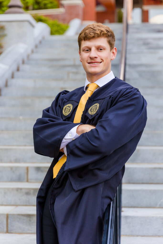 Jason leaning against a stair railing with arms crossed in his graduation gown.