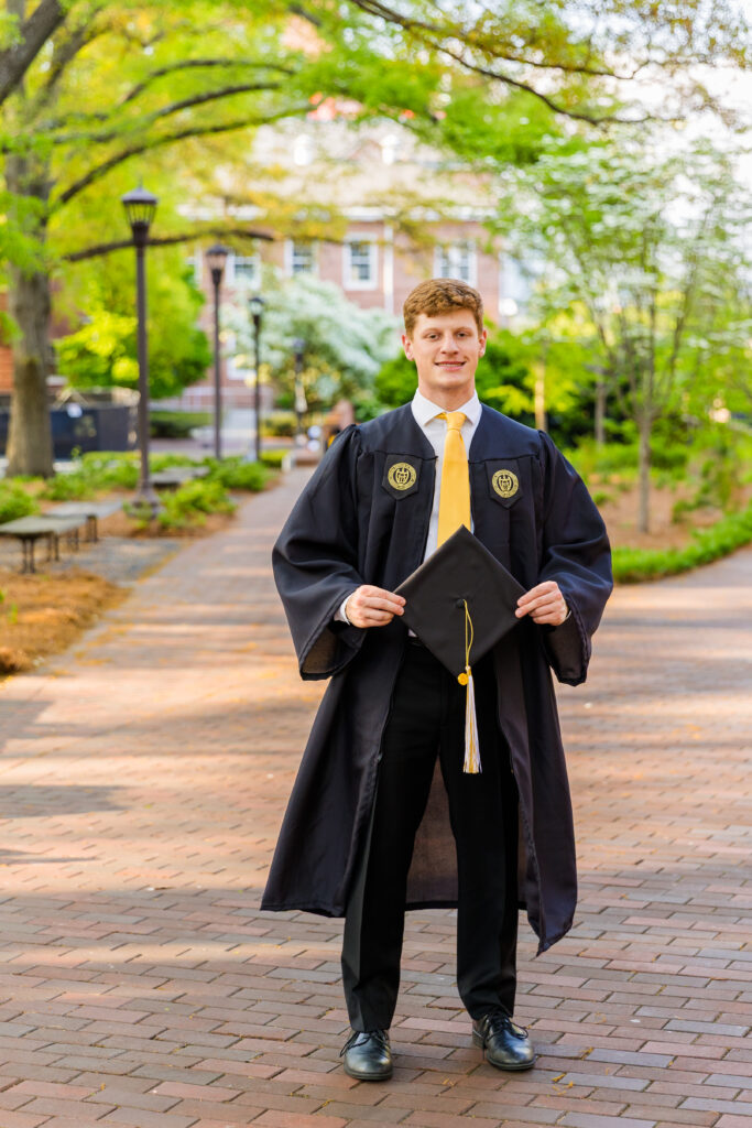 Jason standing on a paved path, holding his graduation cap and facing the camera.