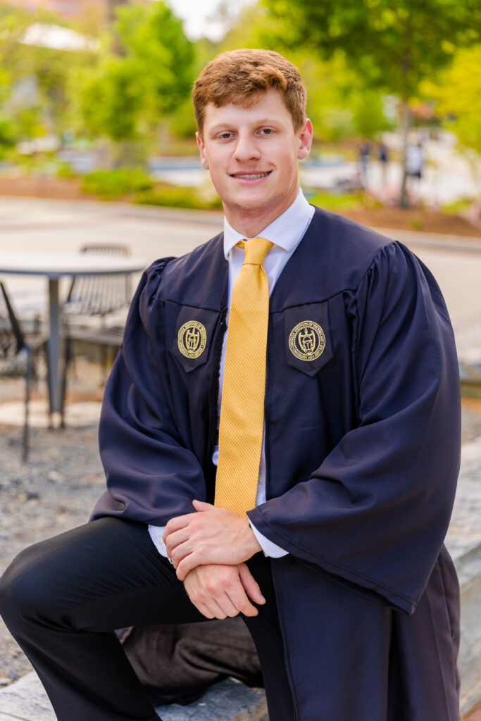 Jason standing casually with one foot raised on a higher step, wearing cap and gown.