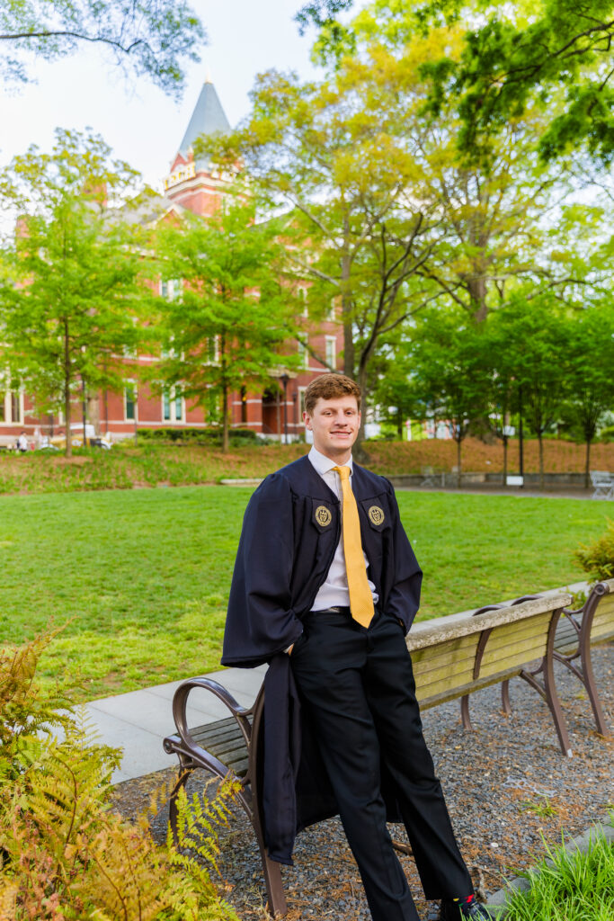 Jason leaning comfortably against the back of a campus bench in his graduation outfit.