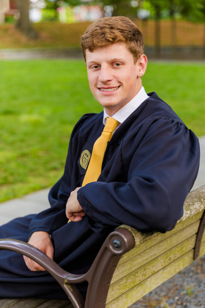 Jason sitting on a bench, looking at the camera over his shoulder, in his graduation outfit.