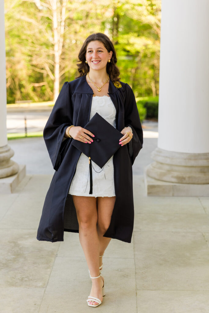 Senior graduate walking towards the camera in her cap and gown, holding the cap during Emory University graduation portraits with Laure Photography.