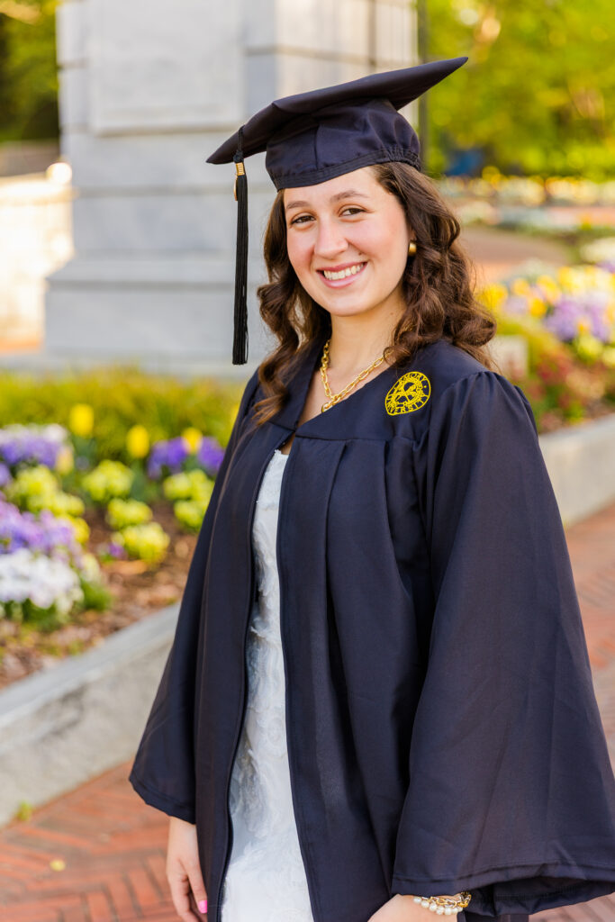 Closeup of senior girl in cap and gown standing in front of Emory gate with flowers, looking sideways during her graduation photo session.