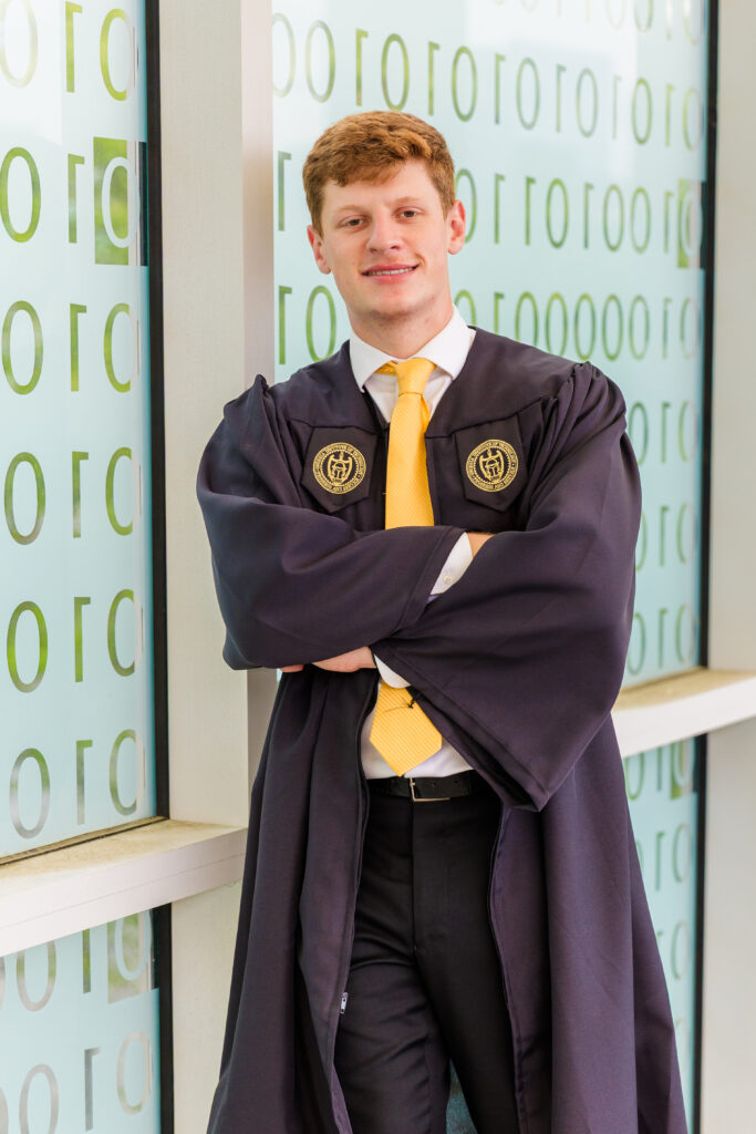 Jason leaning against the transparent binary code wall at Georgia Tech in his graduation outfit.