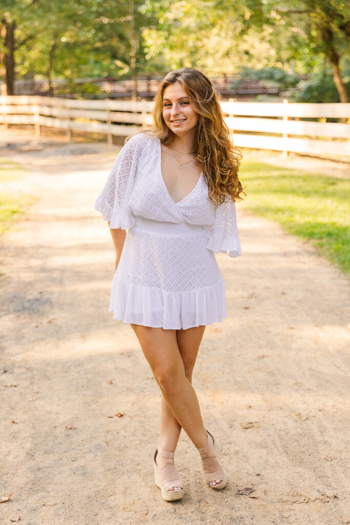 Full-body portrait of Sienna in a white dress standing with hands behind her back and ankles crossed on a path in an Atlanta park