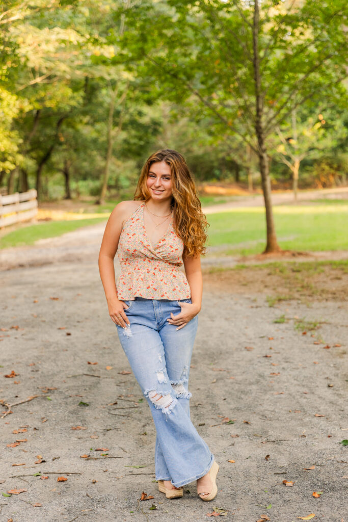 Full-body shot of high school senior wearing jeans and a sleeveless floral top, standing with ankles crossed in an Atlanta park