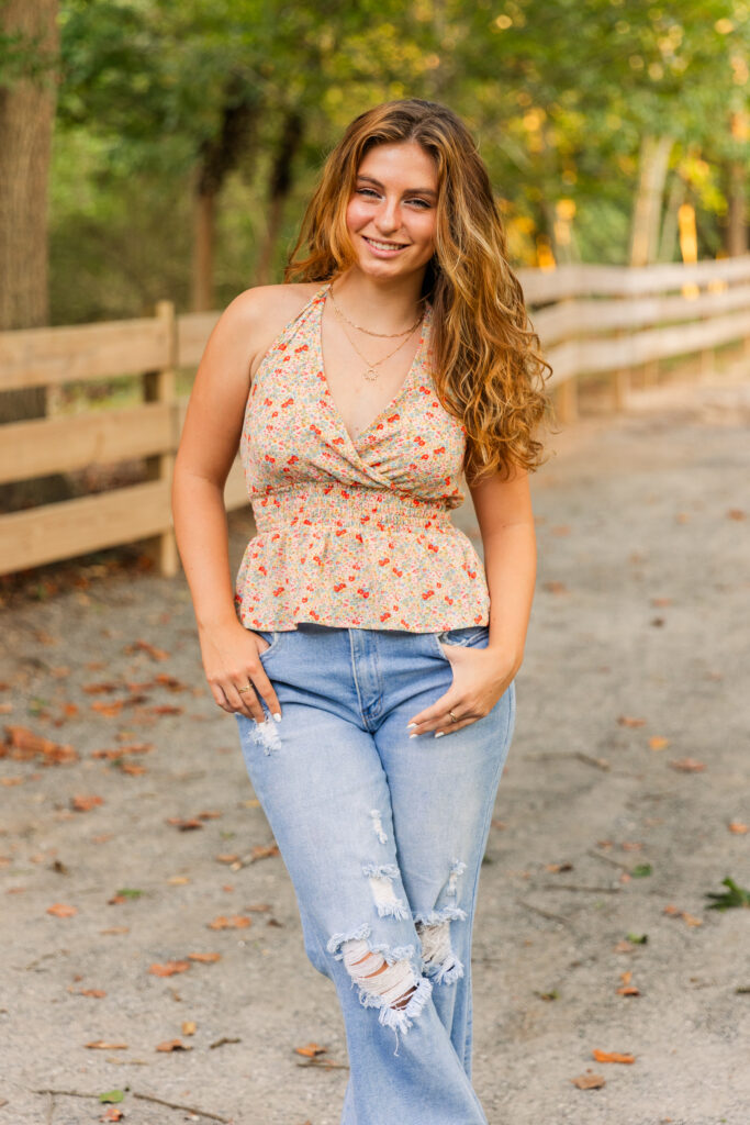 Close-up of high school senior girl wearing a sleeveless floral top and jeans, standing confidently on a park path