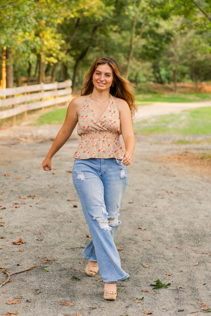 Senior girl confidently walking toward the camera in a floral sleeveless top and jeans on a sunlit park path