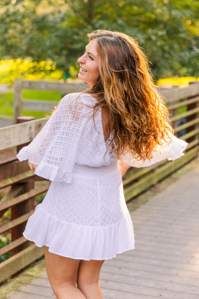 High school senior girl laughing while spinning in a white dress on a wooden bridge during golden hour in an Atlanta park