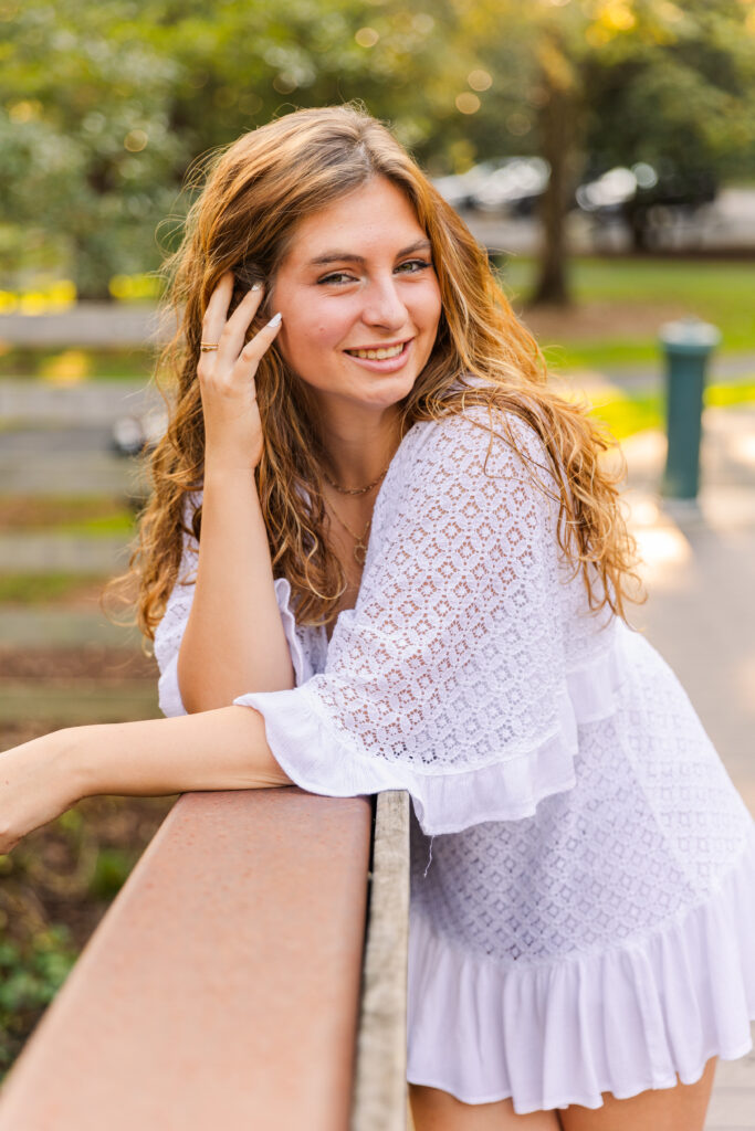 Teen girl in white dress with hand in hair and soft smile, posing on a bridge in Atlanta during senior session