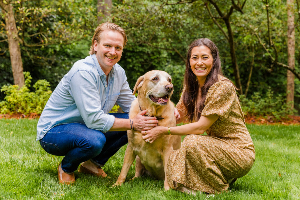 Parents kneeling in the grass with their dog, smiling at the camera.