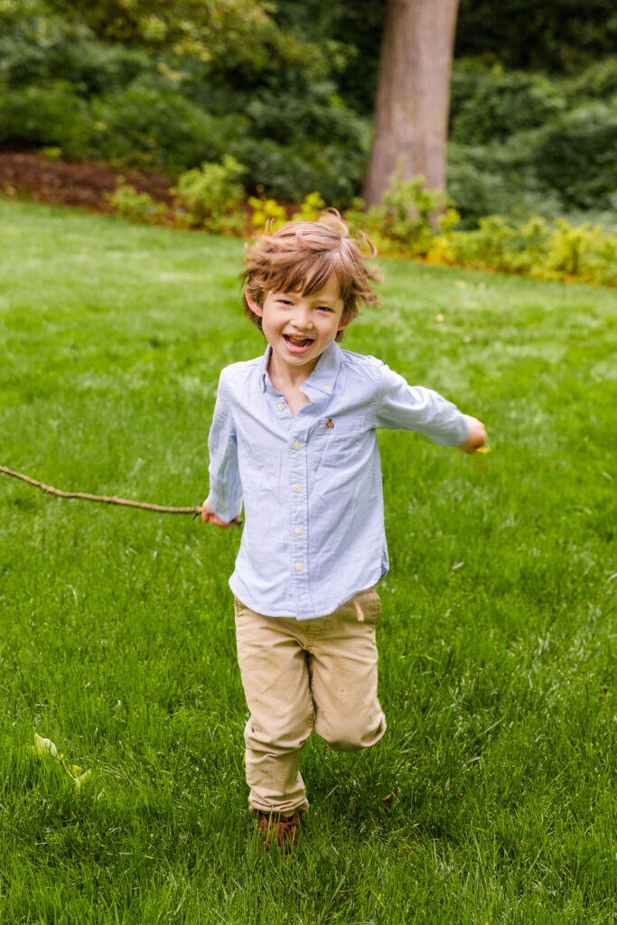 Boy in blue button-down shirt running toward camera holding a stick.