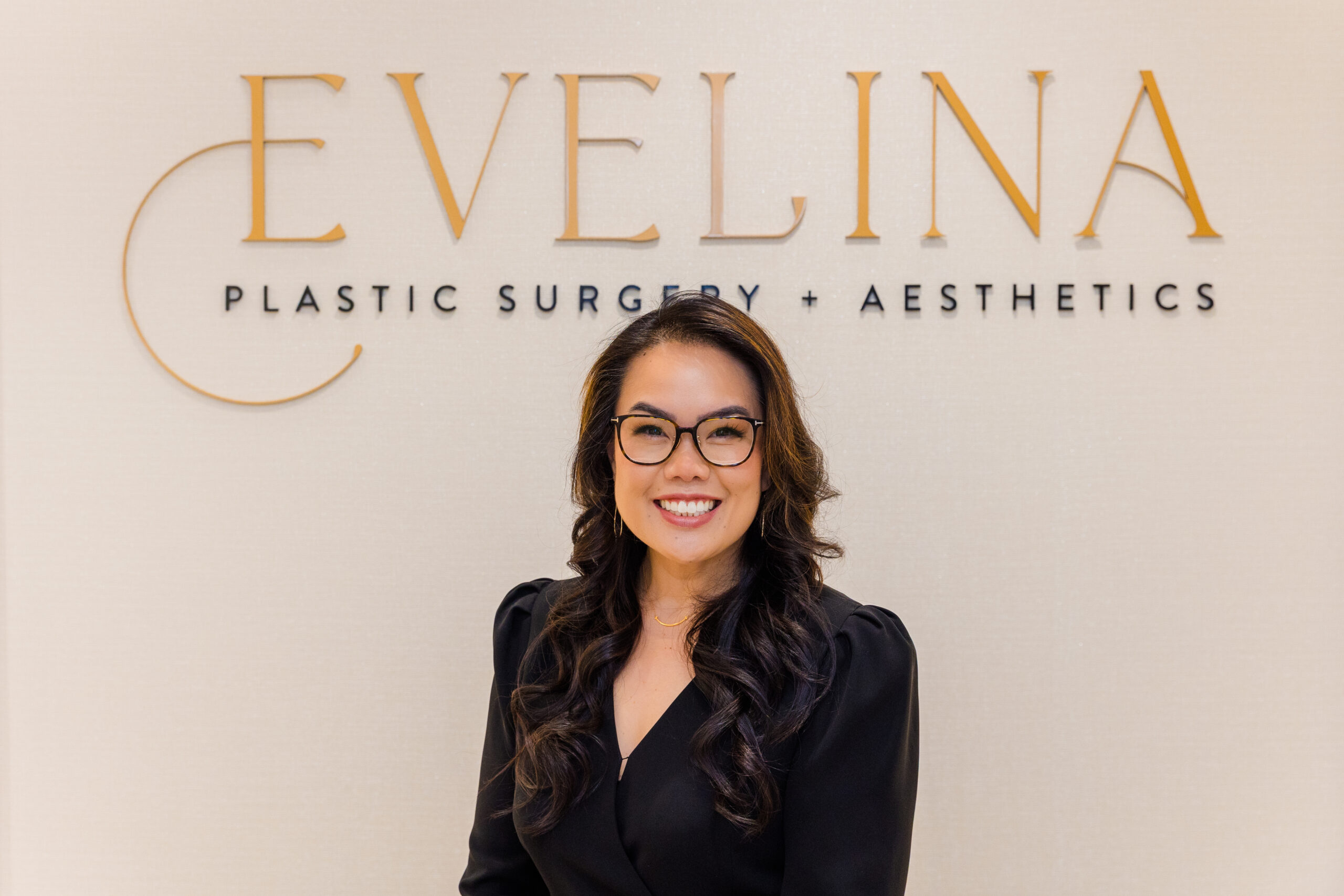 Dr. Katherine Santosa standing under Evelina Plastic Surgery Atlanta wall logo.