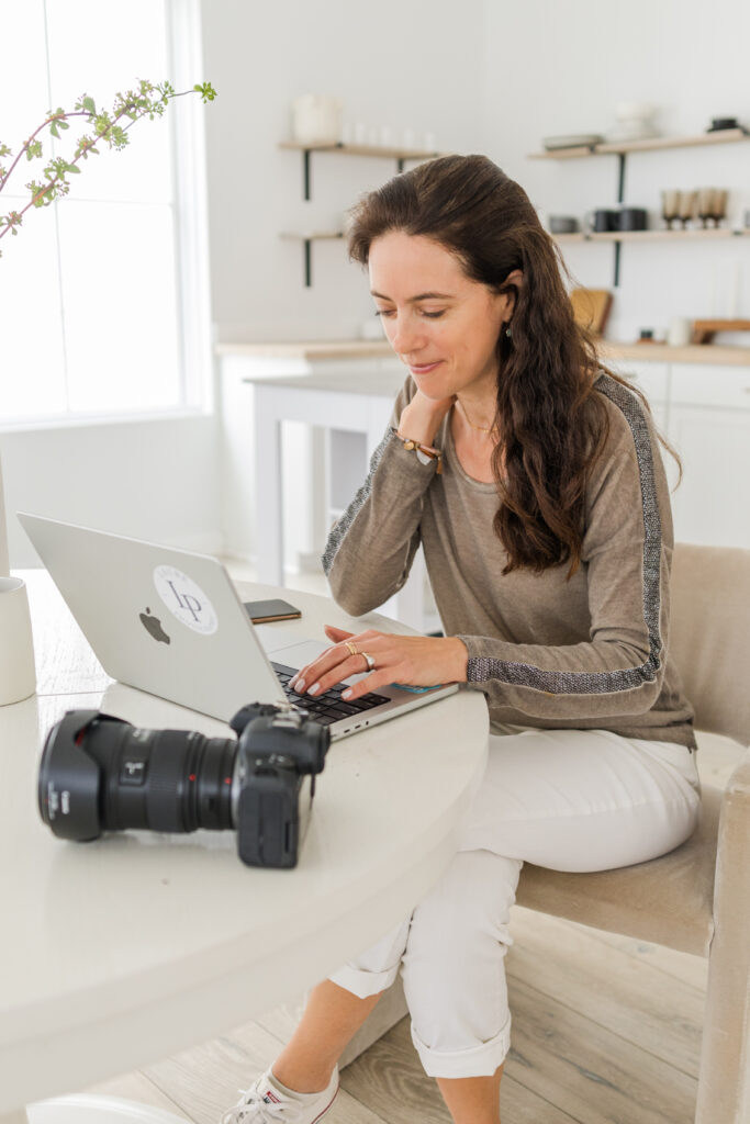woman sitting in her family room working on her computer with her camera next to her with white and beige colors