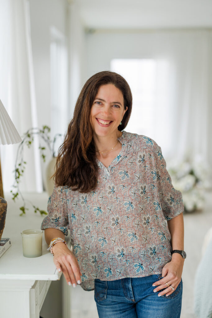 Laure Glorieux standing in a lifestyle environment, one hand resting on a mantelpiece, wearing jeans and a colorful top