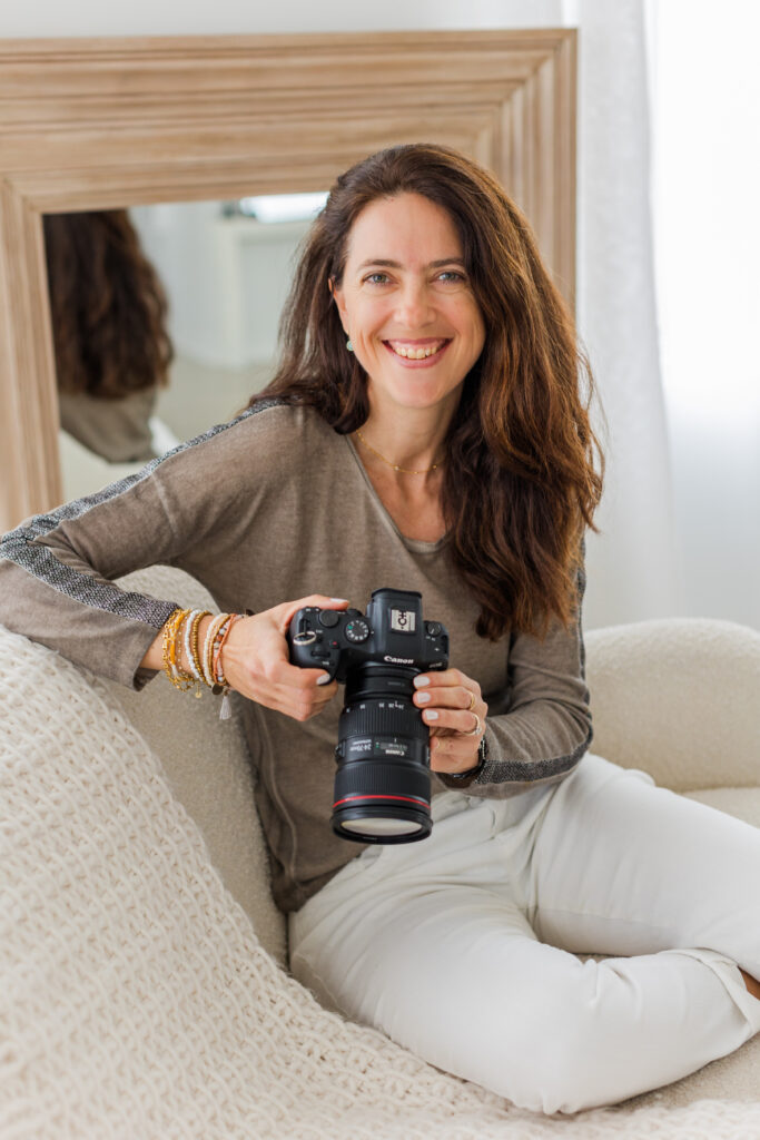 woman wearing white pants and brown top sitting on a beige sofa holding camera and smiling