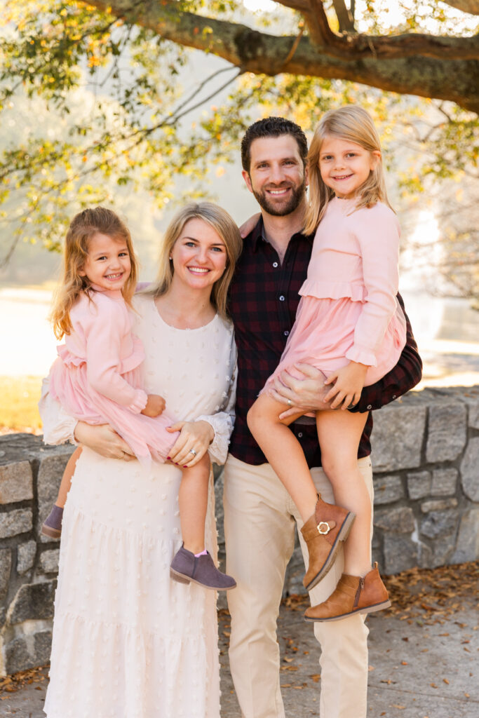 Parents each holding one of their daughters in light pink dresses while sharing a family hug in an Atlanta park during a family photo session with Laure Photography.