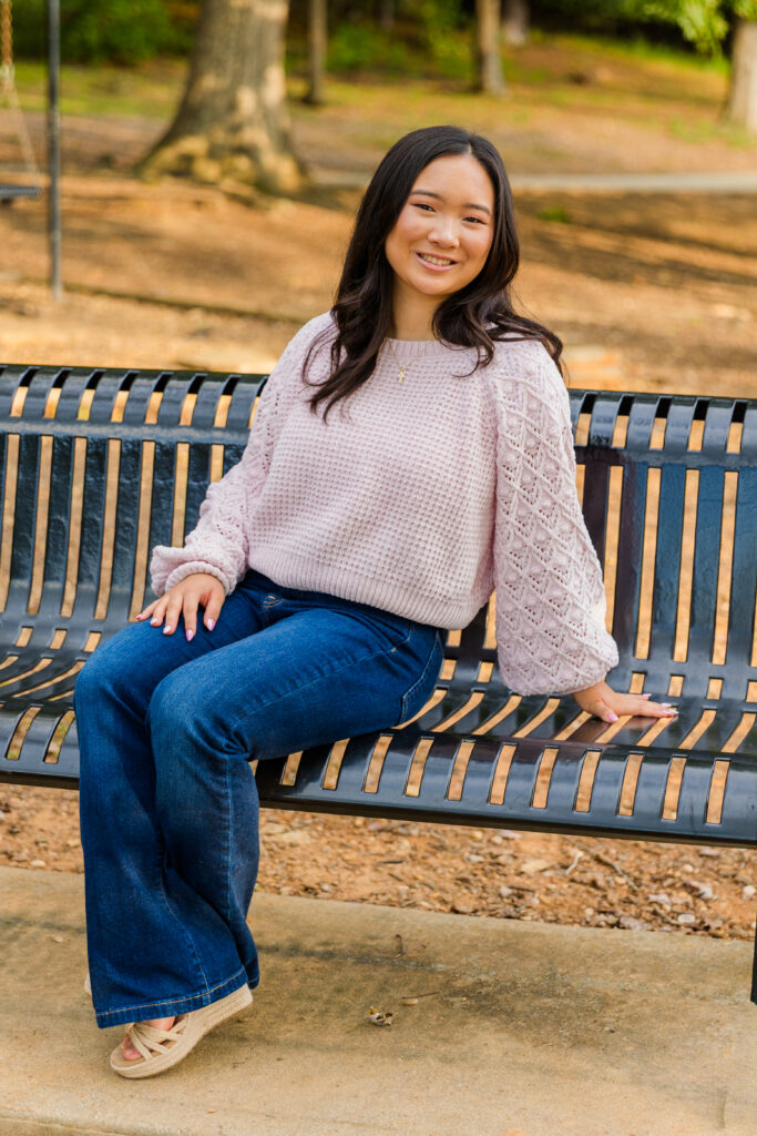 Pace Academy senior sitting on a park bench, ankles crossed, wearing blue jeans and light pink sweater.