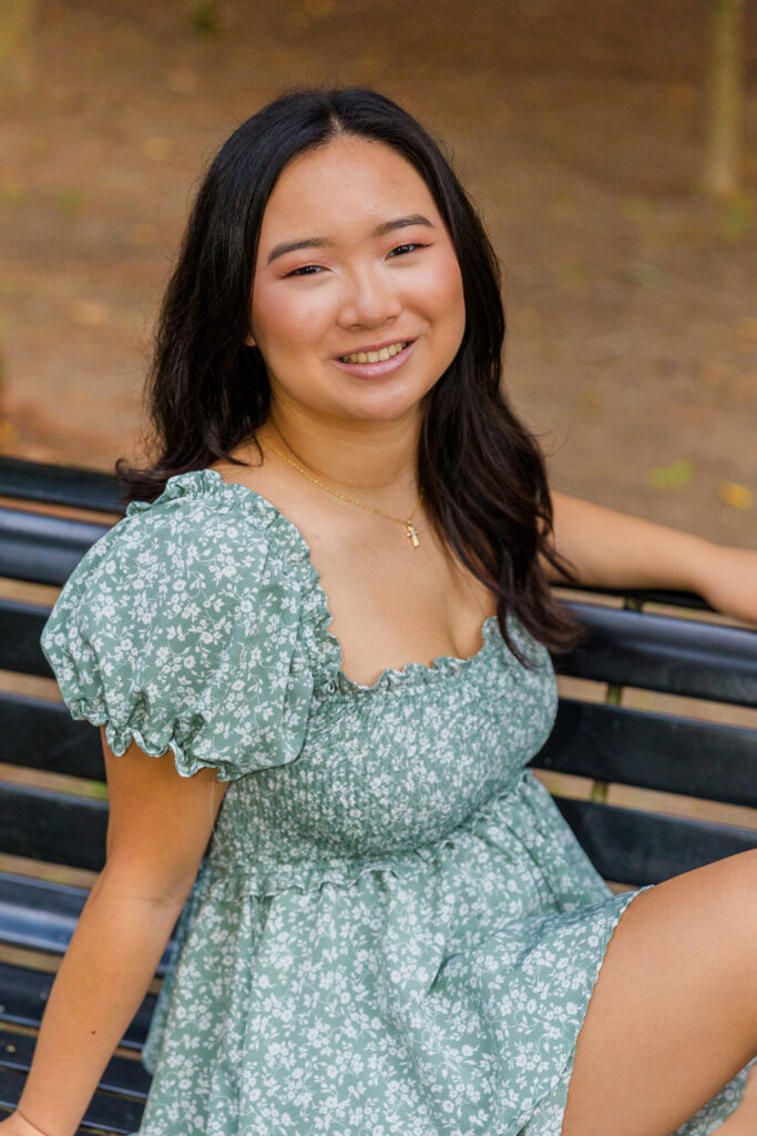 Senior girl in green and white jumper dress sitting sideways on a park bench, one hand resting.