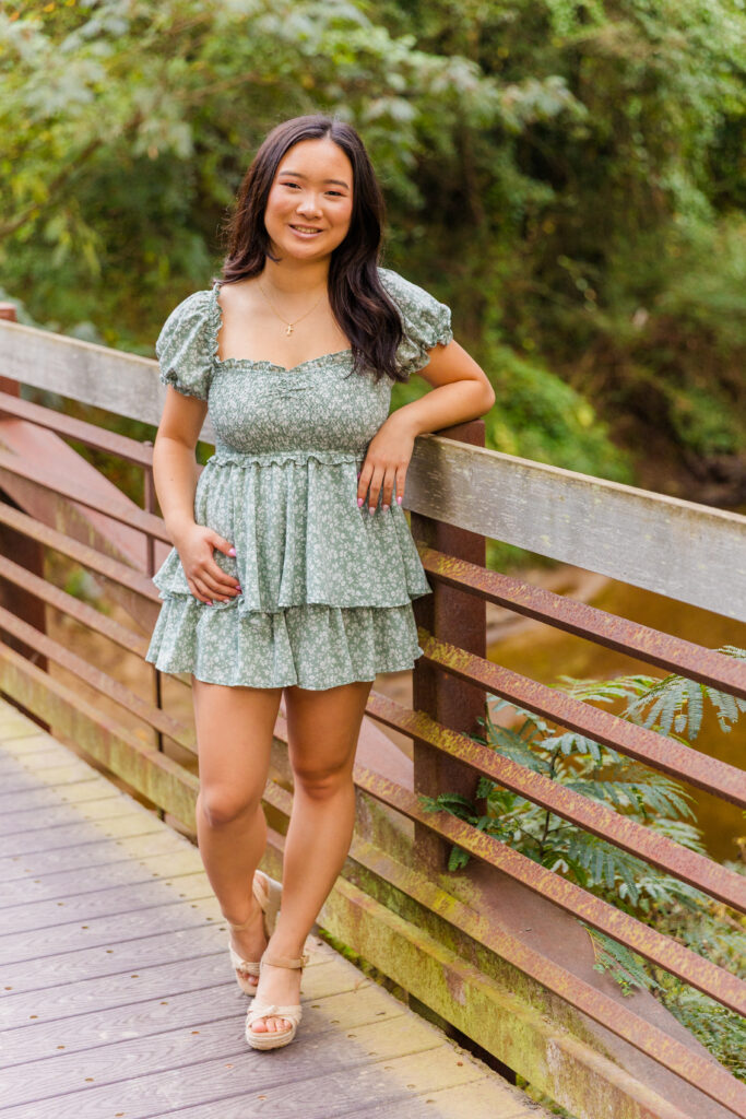 High school senior wearing green and white jumper dress with floral pattern standing on bridge in Brookhaven.