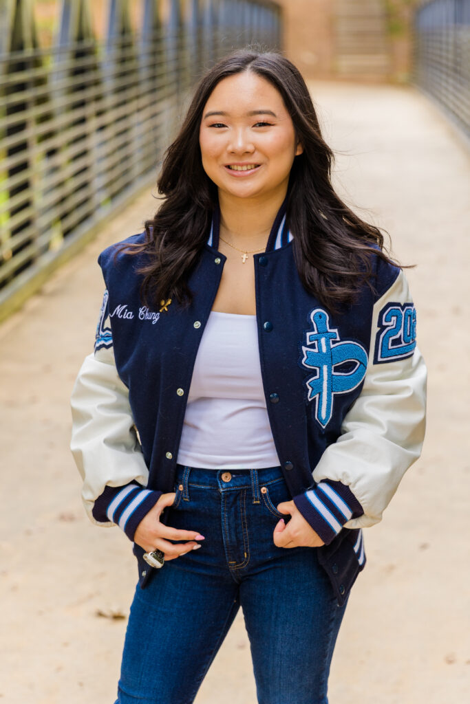 Atlanta senior portrait of Mia in jeans and school jacket standing on a bridge, hands in pockets, smiling.