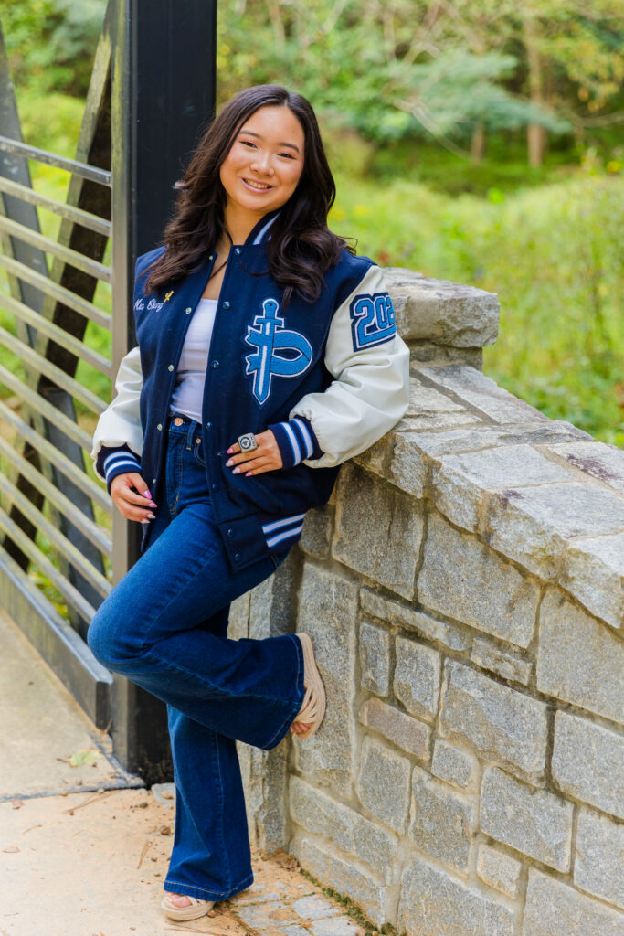 Mia leaning against a bridge pillar in Brookhaven wearing jeans and a leather jacket, one knee up.