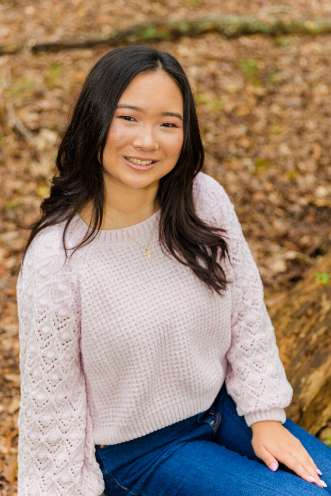 Close-up of Mia sitting on a tree log in Brookhaven park wearing blue jeans and pink sweater.